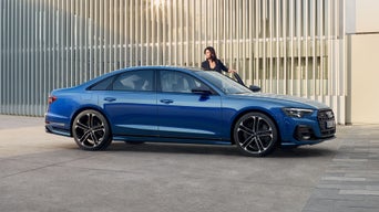 Blauer Audi S8 steht vor einer Wand mit vertikalen Metalllamellen, dunkelhaarige Frau steigt ein.