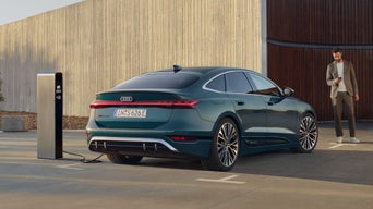 Der S6 Sportback e-tron im schrägen Heckprofil. Das Fahrzeug steht vor einem großen Gebäude mit Holzvertäfelung und ist an eine Ladestation angeschlossen. Vor dem Fahrzeug steht ein junger Mann und blickt auf das Smartphone in seiner Hand.