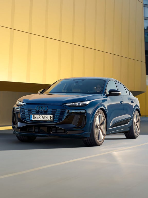 Blauer Audi SQ6 Sportback e-tron fährt auf Straße vor gelber Gebäudefassade, von vorn links fotografiert.