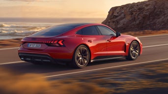 Dynamische Seitenansicht eines roten Audi e-tron GT quattro, der auf einer Küstenstraße fährt. Im Hintergrund sind Klippen, das Meer und der Himmel zu erkennen. Die gesamte Szenerie ist in das Licht eines Sonnenuntergangs gehüllt.
