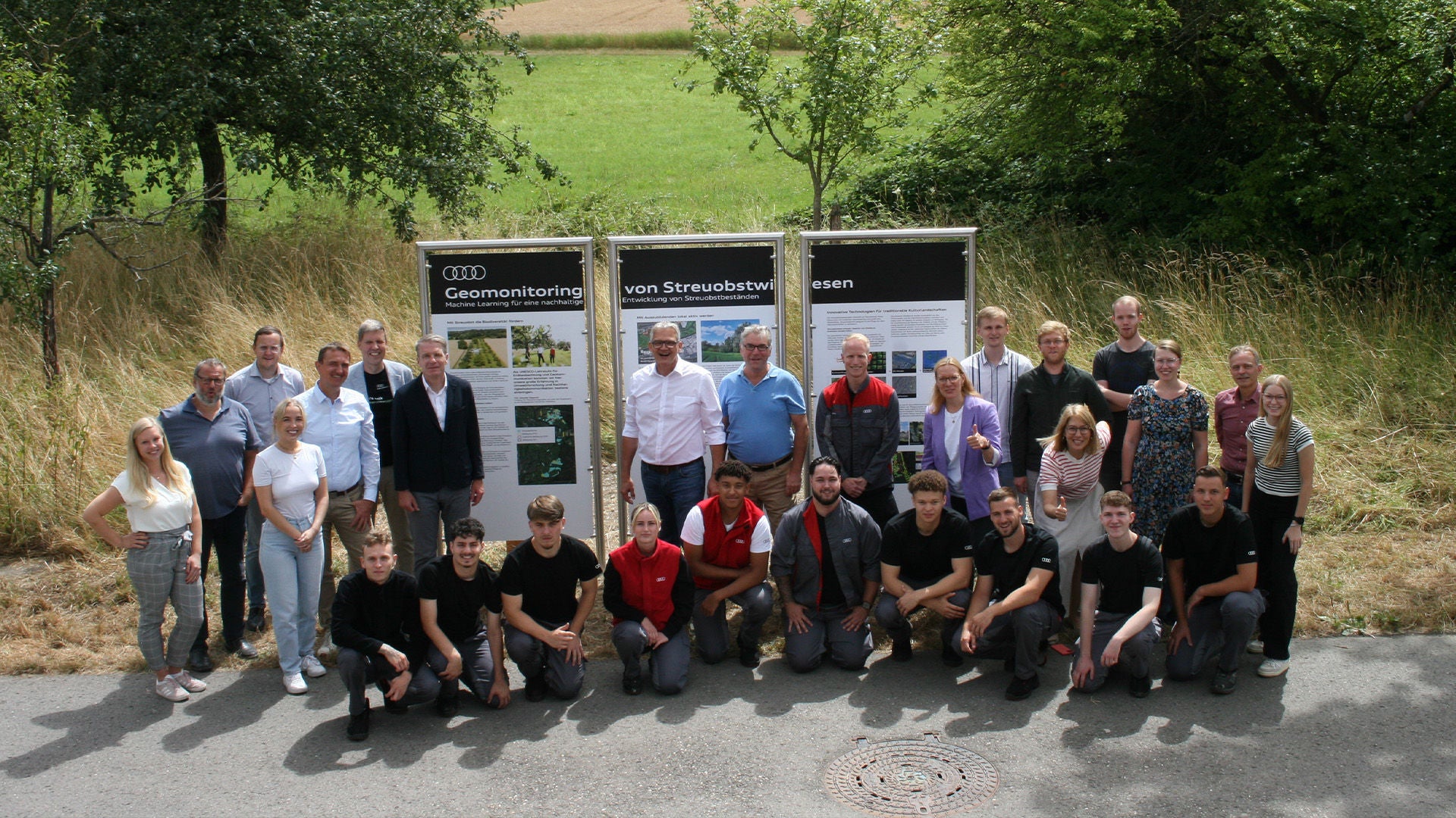 A group of people stand in an orchard next to an information board and look at the camera together.