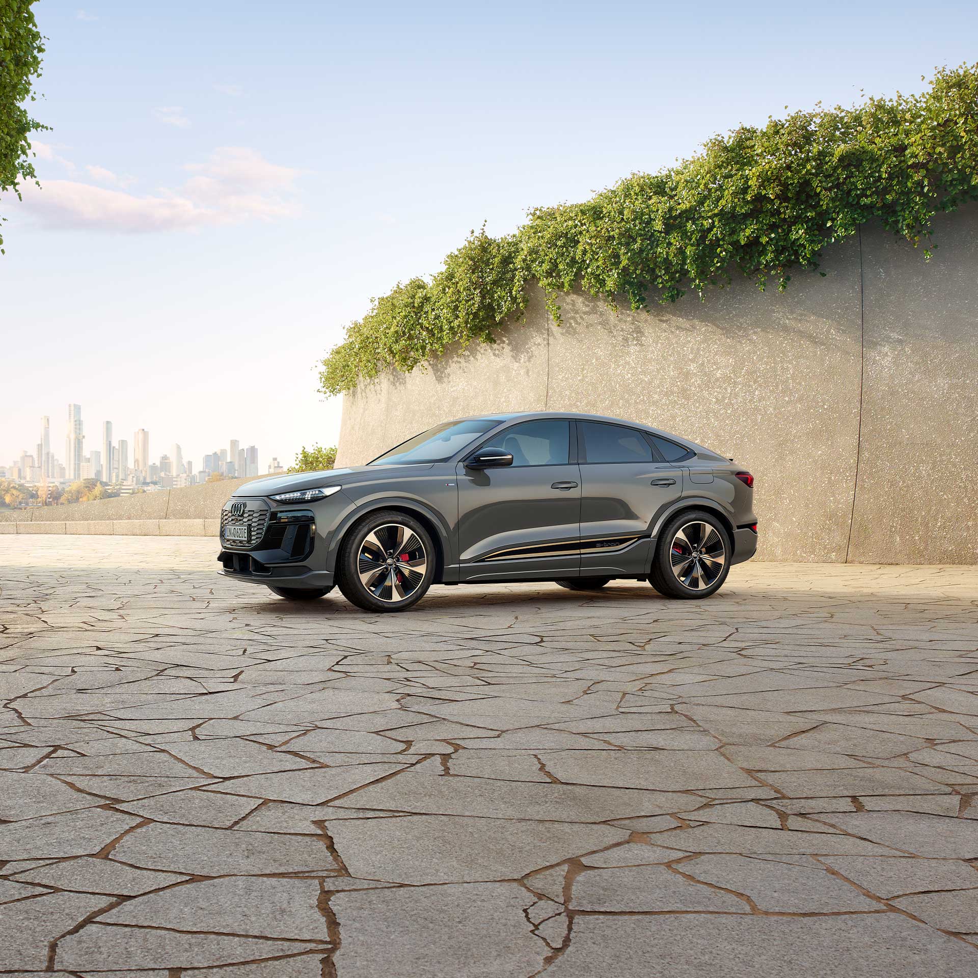 Ein grauer Audi Q6 Sportback e-tron steht auf heller Steinfläche vor einer begrünten Betonwand, Skyline im Hintergrund.