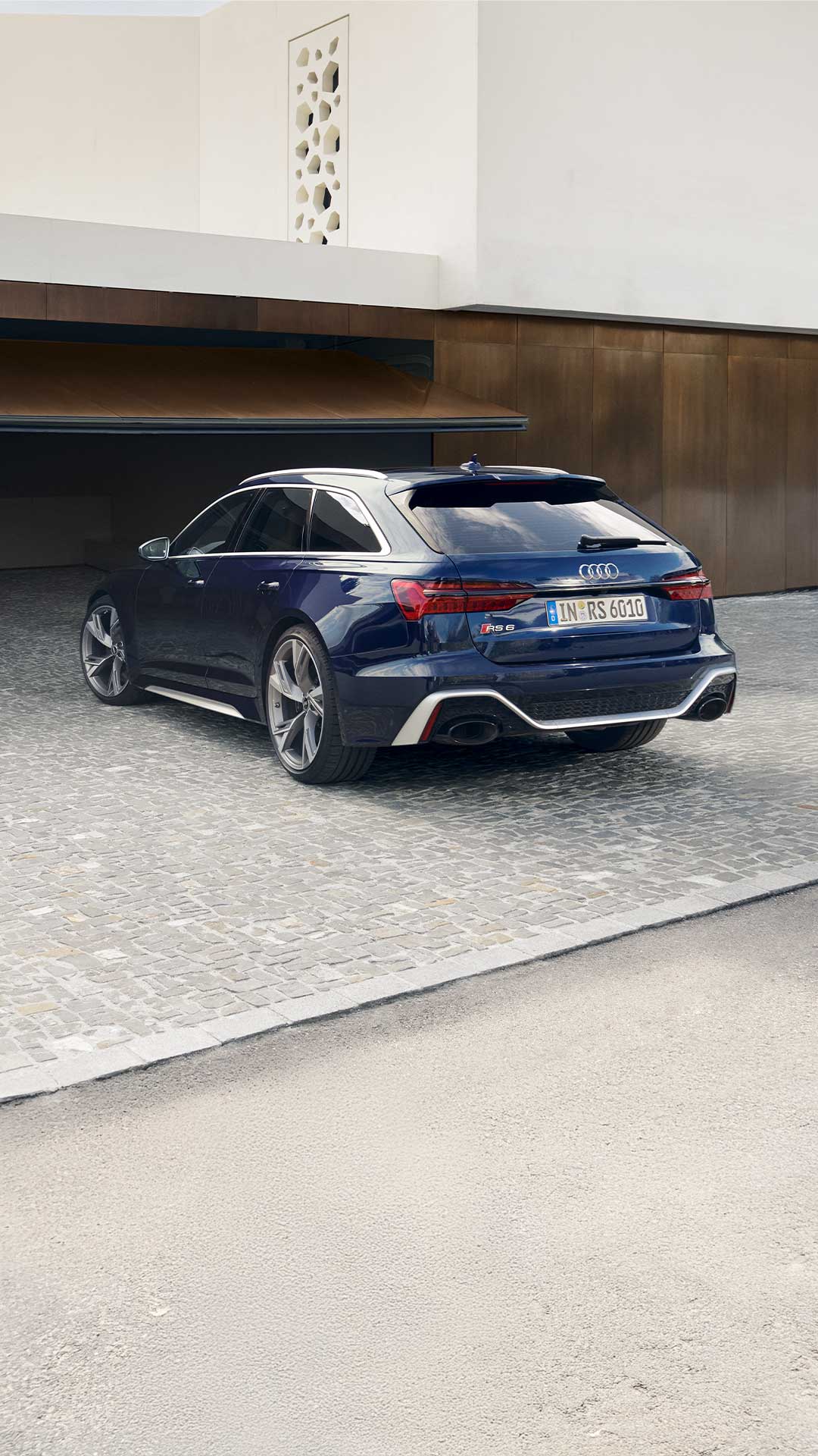 Dunkelblauer Audi RS 6 Avant steht auf gepflasterter Fläche, dunkelhaarige Frau in hellem Business-Outfit läuft rechts am Heck vorbei.