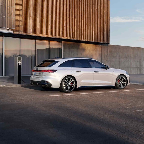 Ein weißer Audi A5 Avant e-hybrid in der Seitenansicht beim Laden vor einer grauen Beton Wand mit großen Fenstern.