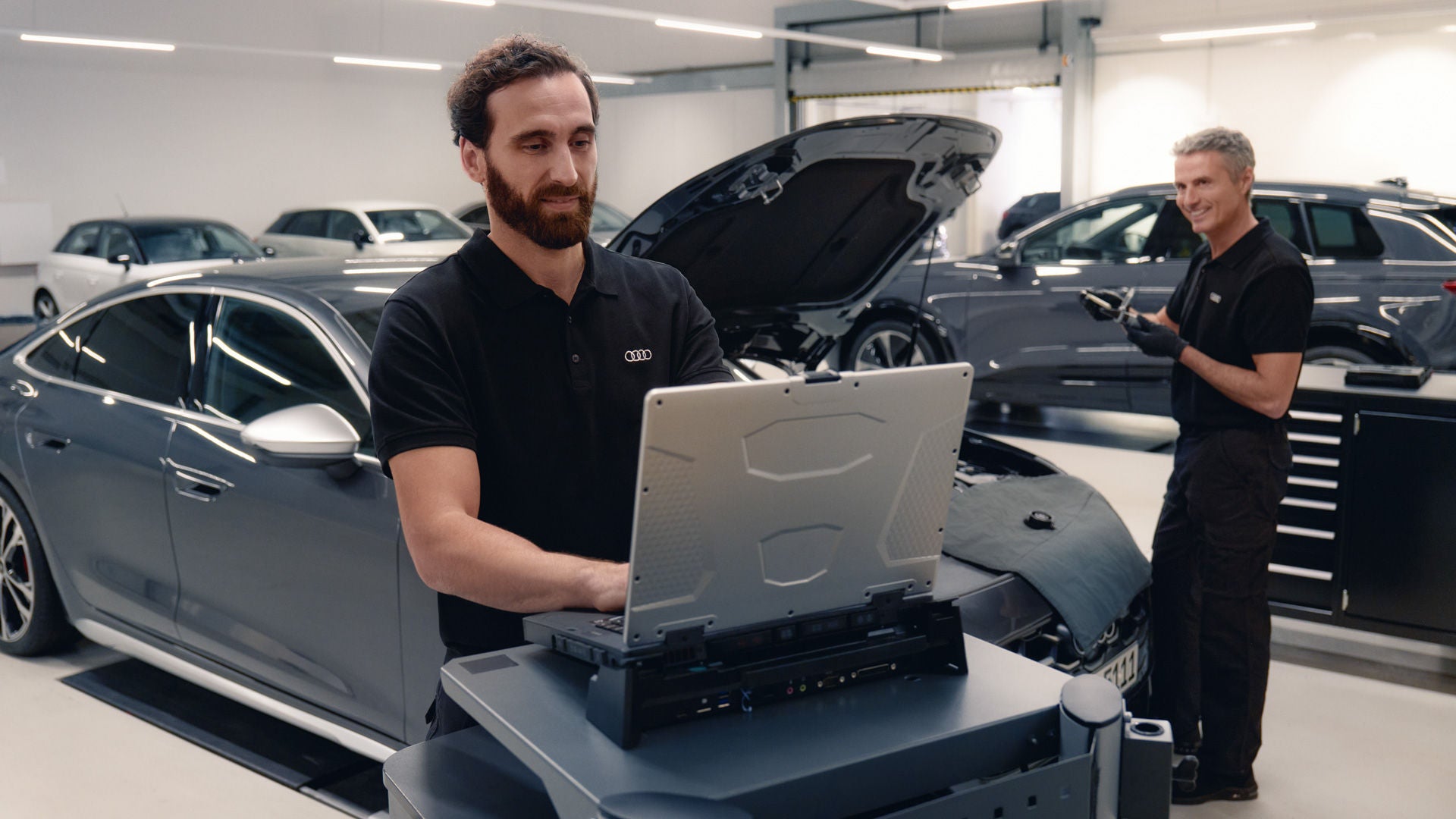 Ein Mann in schwarzer Kleidung steht in einem Autohaus vor einem geöffneten Fahrzeug und bedient ein großes Diagnosegerät. Im Hintergrund steht ein weiterer Mann neben einem Auto.
