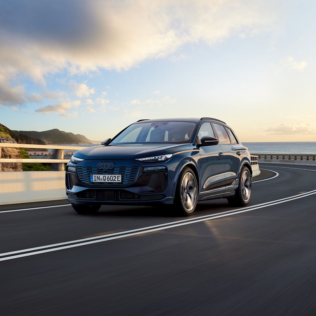 Blauer Audi Q6 e-tron auf einer Küstenstraße bei Sonnenuntergang.