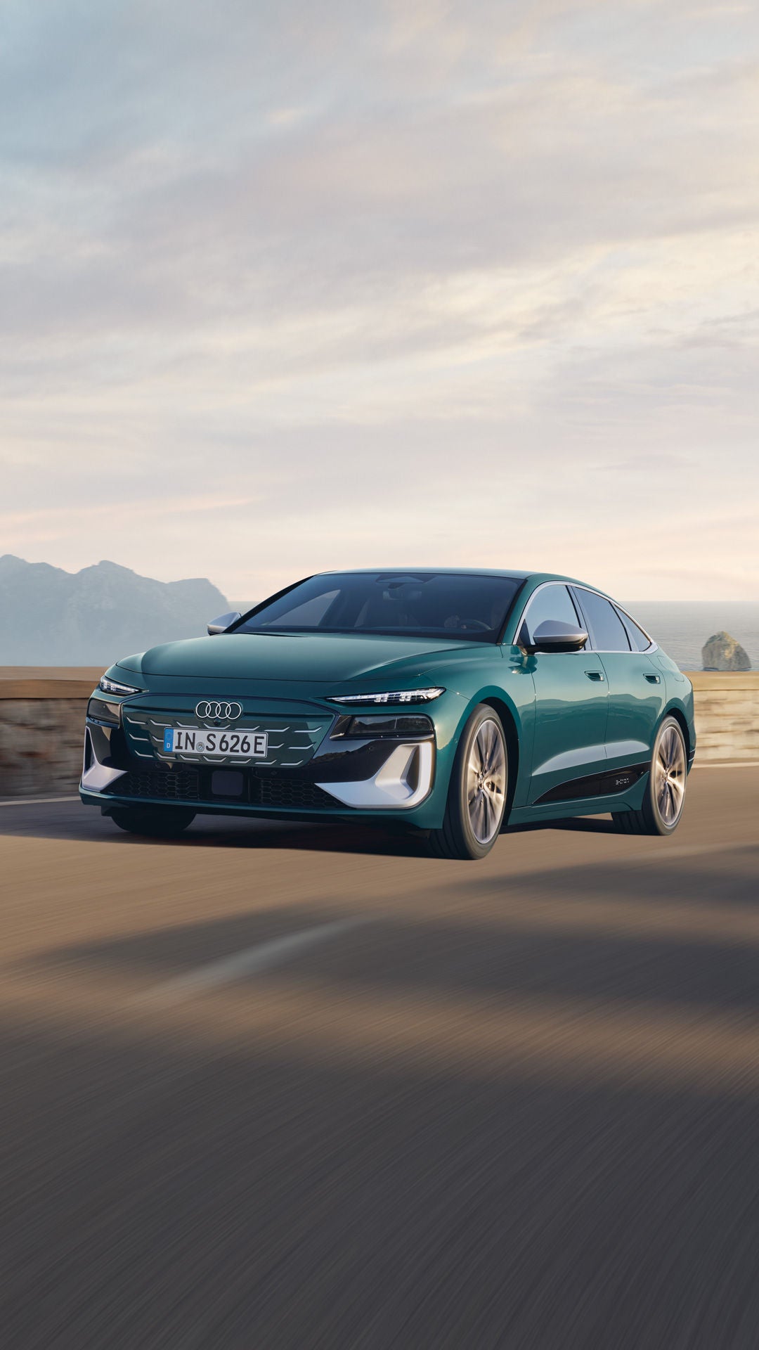 Audi S6 Sportback e-tron in dynamischer Front-Dreiviertelansicht während der Fahrt auf einer Küstenstraße; im Hintergrund sind Meer, Felsen und eine bergige Landschaft zu sehen.
