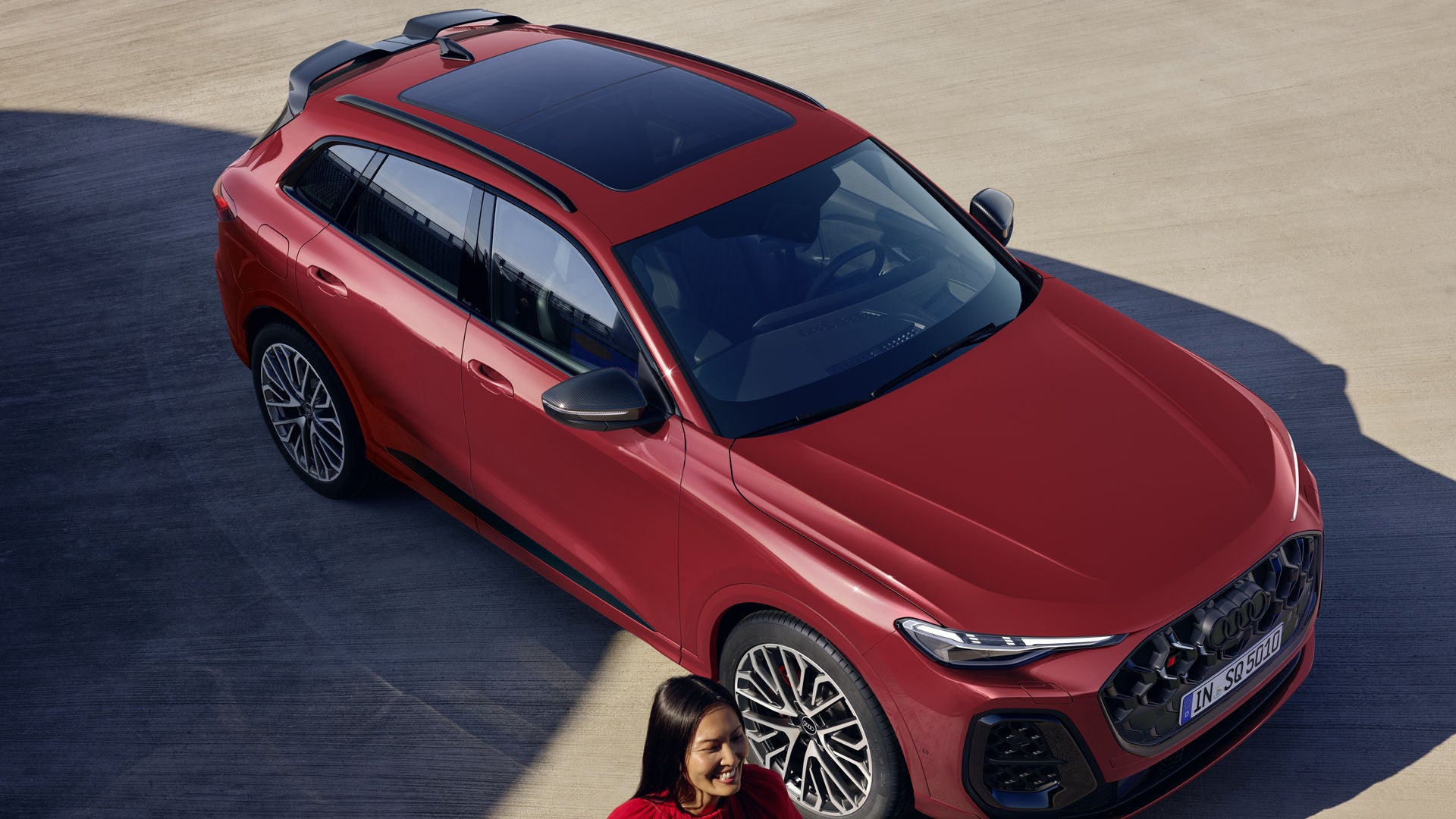 Der Audi SQ5 SUV mit Individuallackierung in Granatrot Perleffekt schräg von oben und vorne. Davor eine modisch gekleidete Frau, die lächelt.