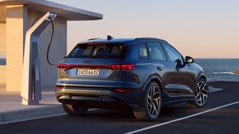 Schräge Heckaufaufnahme des Q6 SUV e-tron, welcher an eine Ladestation auf einem Parkplatz angeschlossen ist. Im Hintergrund ist das Meer zu sehen.