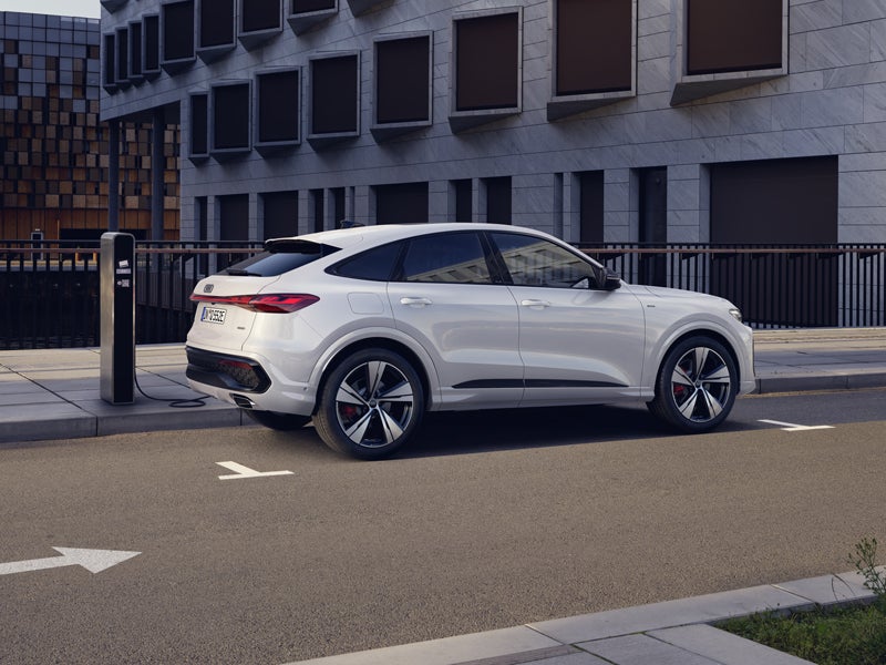 Seitenprofil eines weißen Q5 Sportback e-hybrid, der auf dem Parkplatz vor einer Hochhausfassade lädt.&nbsp;
