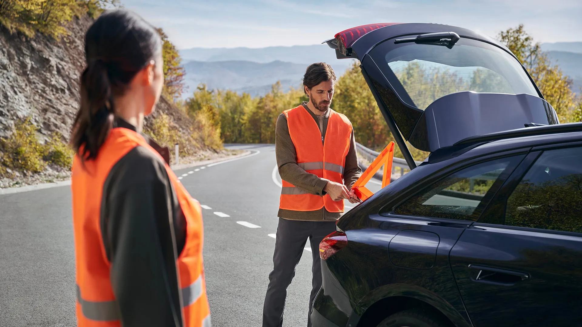 Audi driver roadside assistance scenario on a rural mountain road. A vehicle is parked with the tailgate open while two people wearing high-visibility orange vests stand beside it. One person removes a warning triangle from the luggage compartment, preparing it for use. The winding road is surrounded by trees, rocky hillsides and distant mountains under clear daylight conditions.