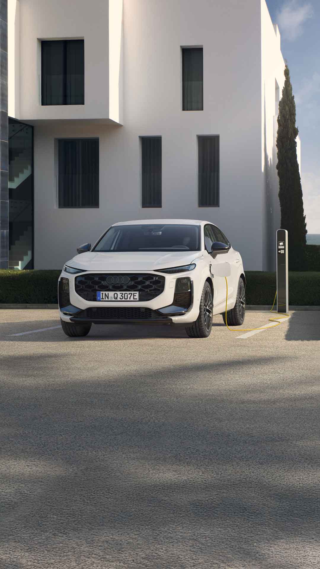 Audi Q3 Sportback e-hübriid külgvaates tagantvaates heledate kividega parklas. Auto laeb moodsas laadimisjaamas. Auto ees on lühikeste juustega naine, kes autost eemale kõnnib.