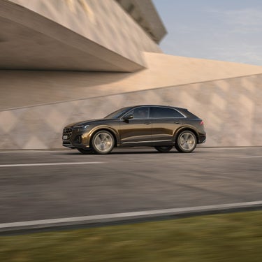 Ein brauner Audi Q8 SUV TFSI e fährt auf einer Straße vor einer strukturierten Wand bei Tageslicht.