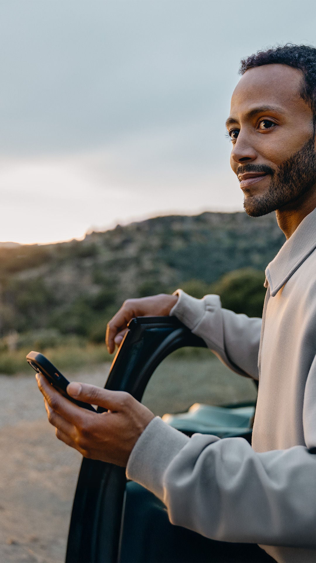 A man with a mobile phone in his hand leans gently smiling against the open driver's door of a vehicle in front of a wooded mountain backdrop at sunrise.