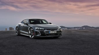Grauer Audi-Sportwagen steht vor städtischer Kulisse auf freier Asphaltfläche bei Sonnenuntergang.