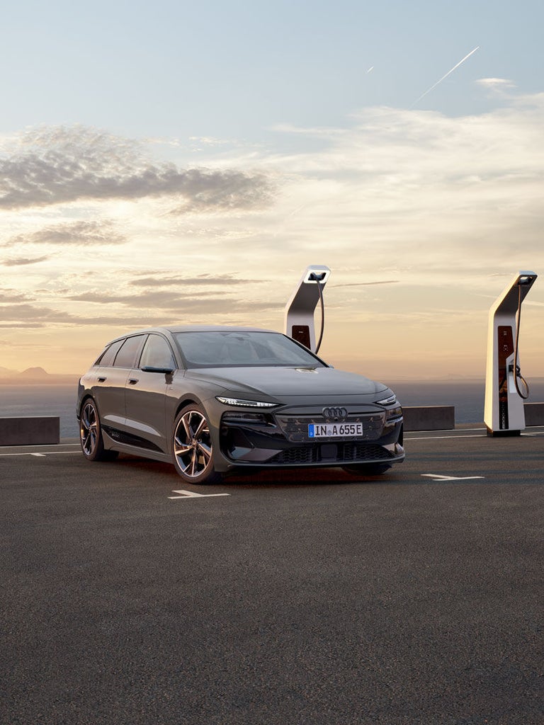 Grauer Audi A6 Avant e-tron steht an Ladesäule auf Küstenparkplatz bei Sonnenuntergang, Mann blickt aufs Meer.