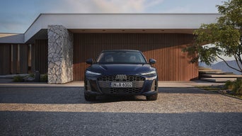 A black Audi is parked in front of a modern house with wooden paneling and stone accents, under a clear sky, conveying luxury and elegance.