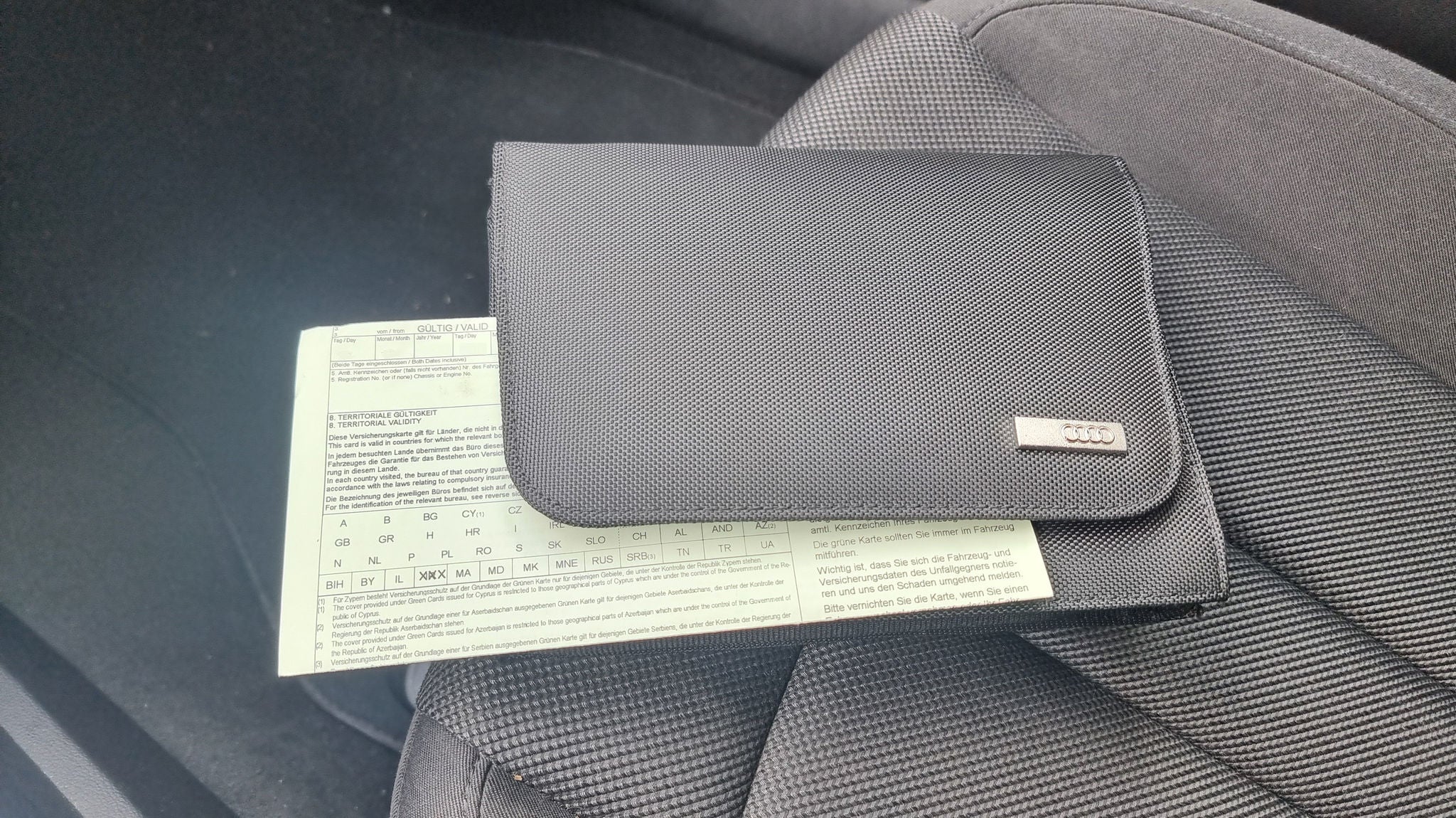 Schwarze Audi-Mappe mit sichtbarem Fahrzeugschein liegt auf dem Beifahrersitz eines Autos.