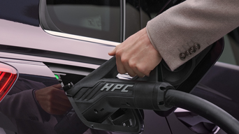 A hand in a beige jacket sleeve holds a black electric car charger with "HPC" text, connected to a vehicle's charge port.