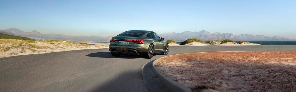 Grüner Audi RS e-tron GT performance fährt auf einer Küstenstraße mit Dünen, Meer und Bergen im Hintergrund.