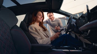 Frau sitzt im Fahrersitz eines Audi, Mann erklärt Funktionen am geöffneten Fahrzeug.