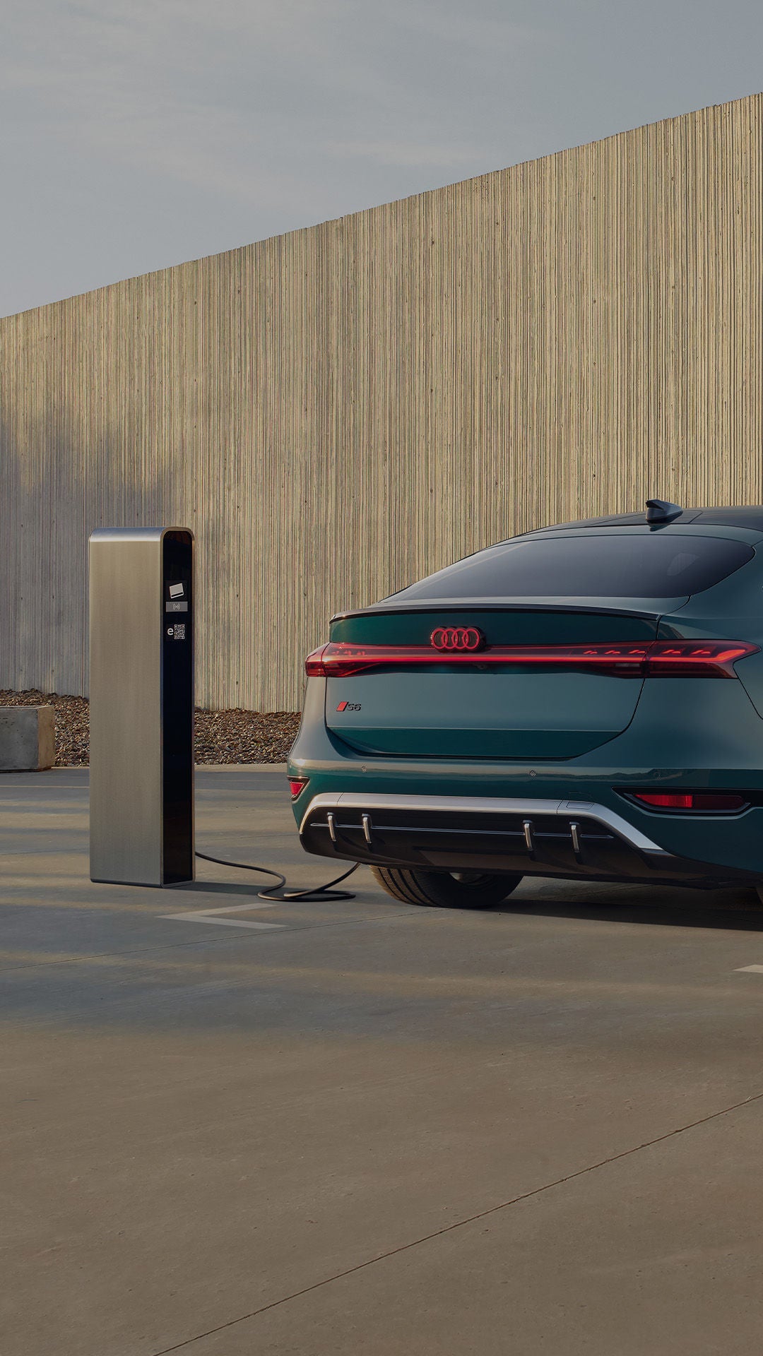 An Audi electric car is parked and charging at a charging station, surrounded by trees and modern concrete structures.
