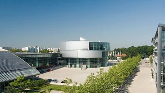 Moderne Glas- und Stahlarchitektur des Gebäudes „museum mobile“ bei Tageslicht, umgeben von Bäumen und Gehweg.