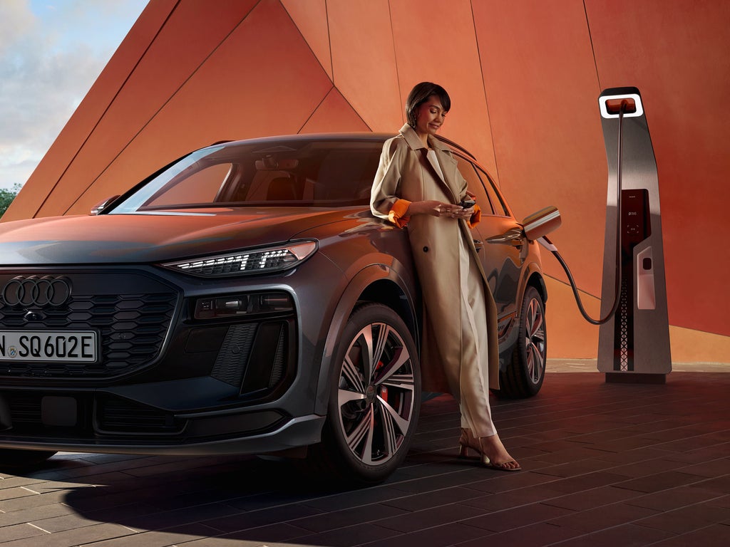 Der SQ6 SUV e-tron steht vor einer orangenen Gebäudefassade und ist dabei an eine Ladestation angeschlossen. An der Seite des Fahrzeugs lehnt sich eine junge Frau an und schaut auf ihr Handy. 