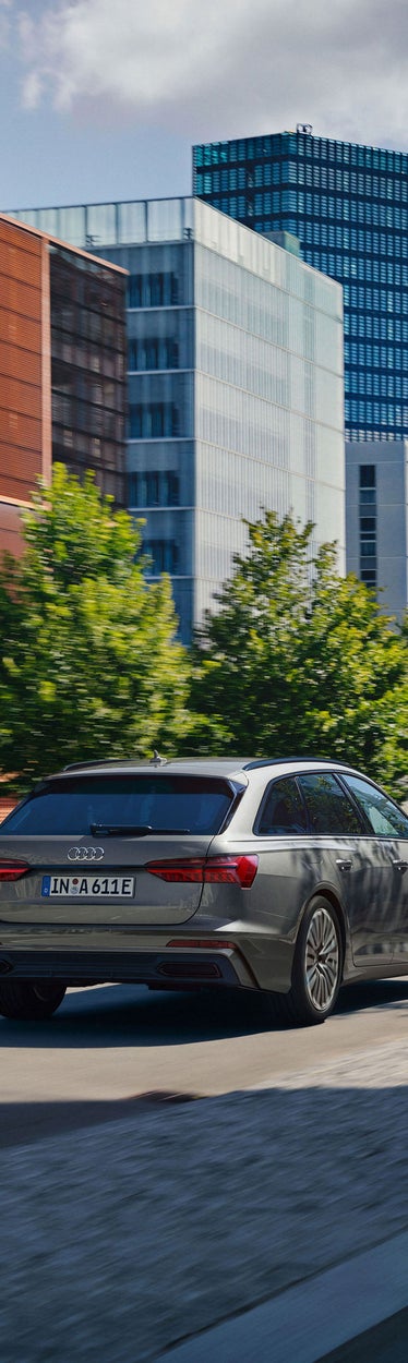 Ein grauer Audi A6 Avant TFSI e fährt durch eine moderne Stadtstraße mit Bäumen und Bürogebäuden im Hintergrund.
