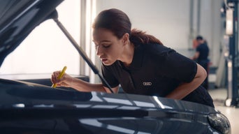 Frau im Audi-Poloshirt markiert mit gelbem Stift eine Stelle am Fahrzeug in einer Werkstatt.