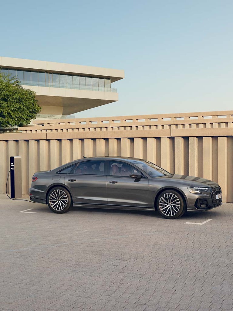 Seitenansicht eines grauen Audi A8 L 60 TFSI e an Ladesäule vor Betonwand, Fahrzeug wird über Fahrerseite geladen.