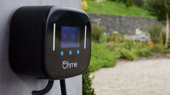 Black electric vehicle charger with digital display and blue buttons mounted on wall, "Ohme" logo, cord hanging down, blurred garden background.
