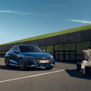 A blue Audi car parked with a person seated beside it, holding a small bouquet, on a sunny day.