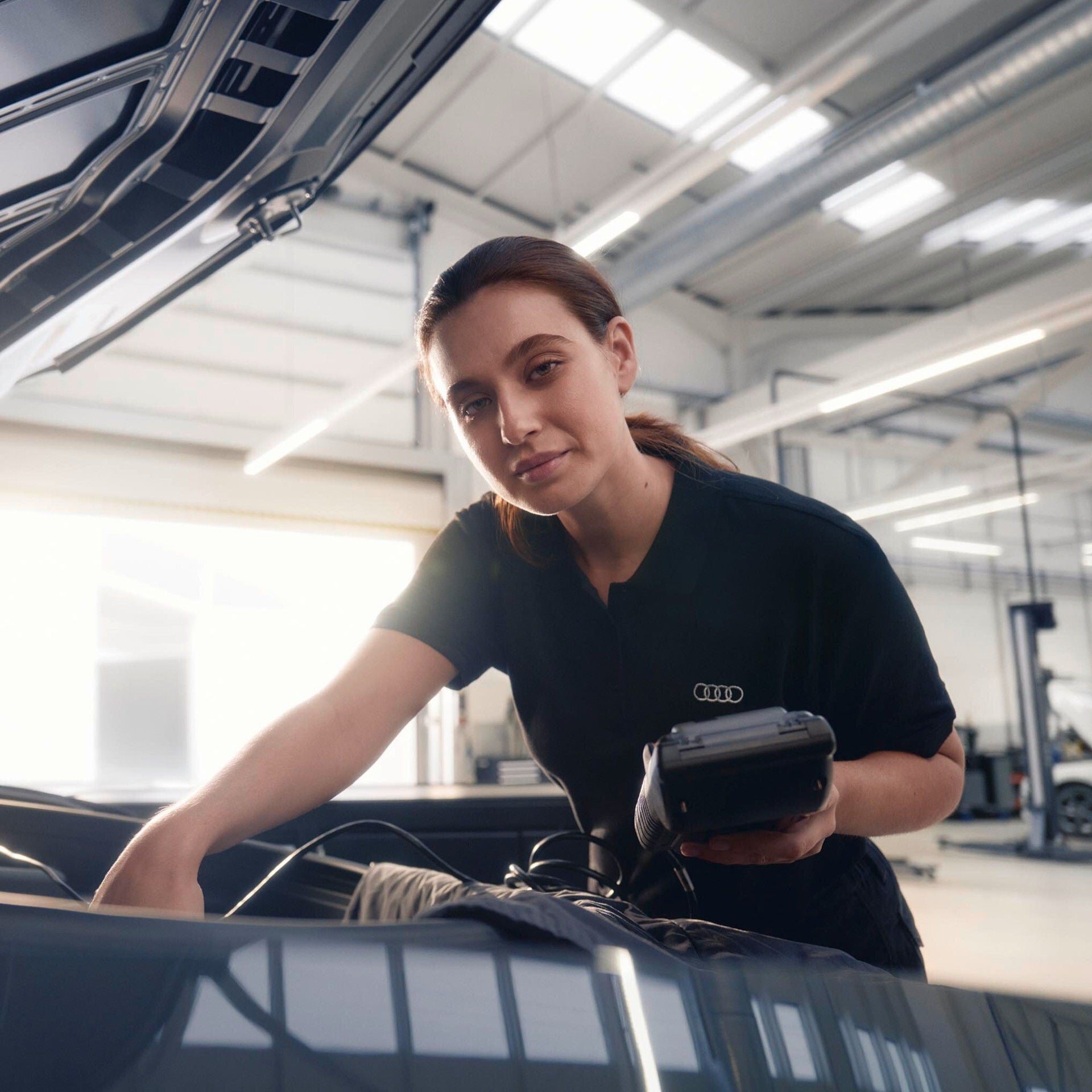Mitarbeiterin prüft im geöffneten Motorraum eines Autos mit einem Diagnosegerät.