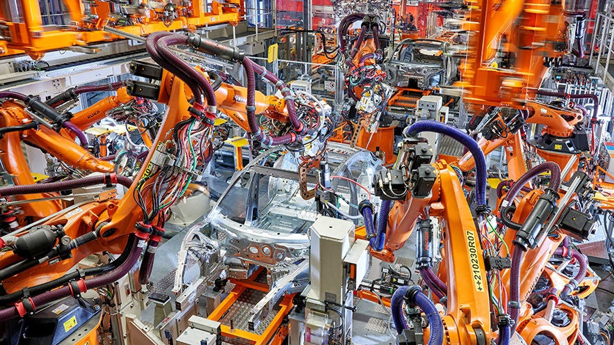 Industrial manufacturing scene with numerous orange robots, cables and a metal vehicle body in a production facility.