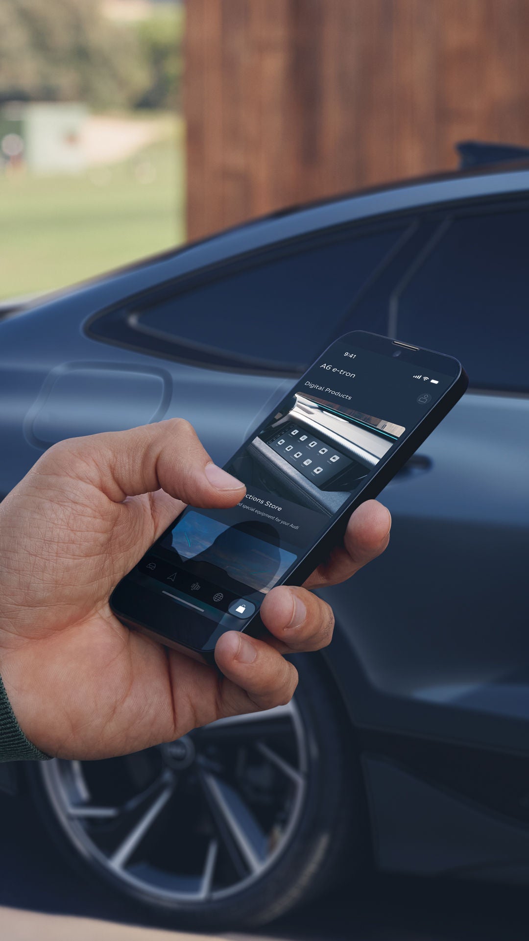 De hand van een man steekt vanaf de linkerkant in het beeld.  Hij houdt een smartphone in zijn hand.  Een donkerblauwe Audi A6 staat iets onscherp op de achtergrond.