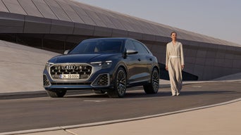 Eine Frau steht neben einem geparkten, dunkelblauen Audi SQ8 SUV vor einem modernen Gebäude.