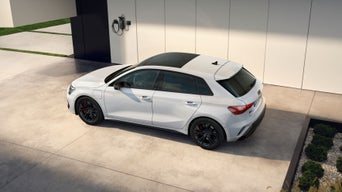White hatchback car parked outside modern house near wall-mounted charging unit.