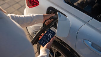 Person lädt weißes Elektroauto und hält ein Smartphone mit Ladeinformationen in der Hand. 