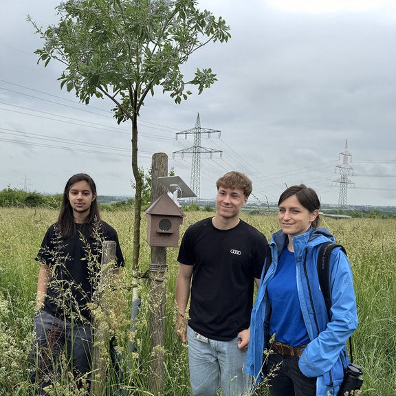 Two teenagers and a woman stand next to a mounted listening box on a wooden post in a meadow landscape.