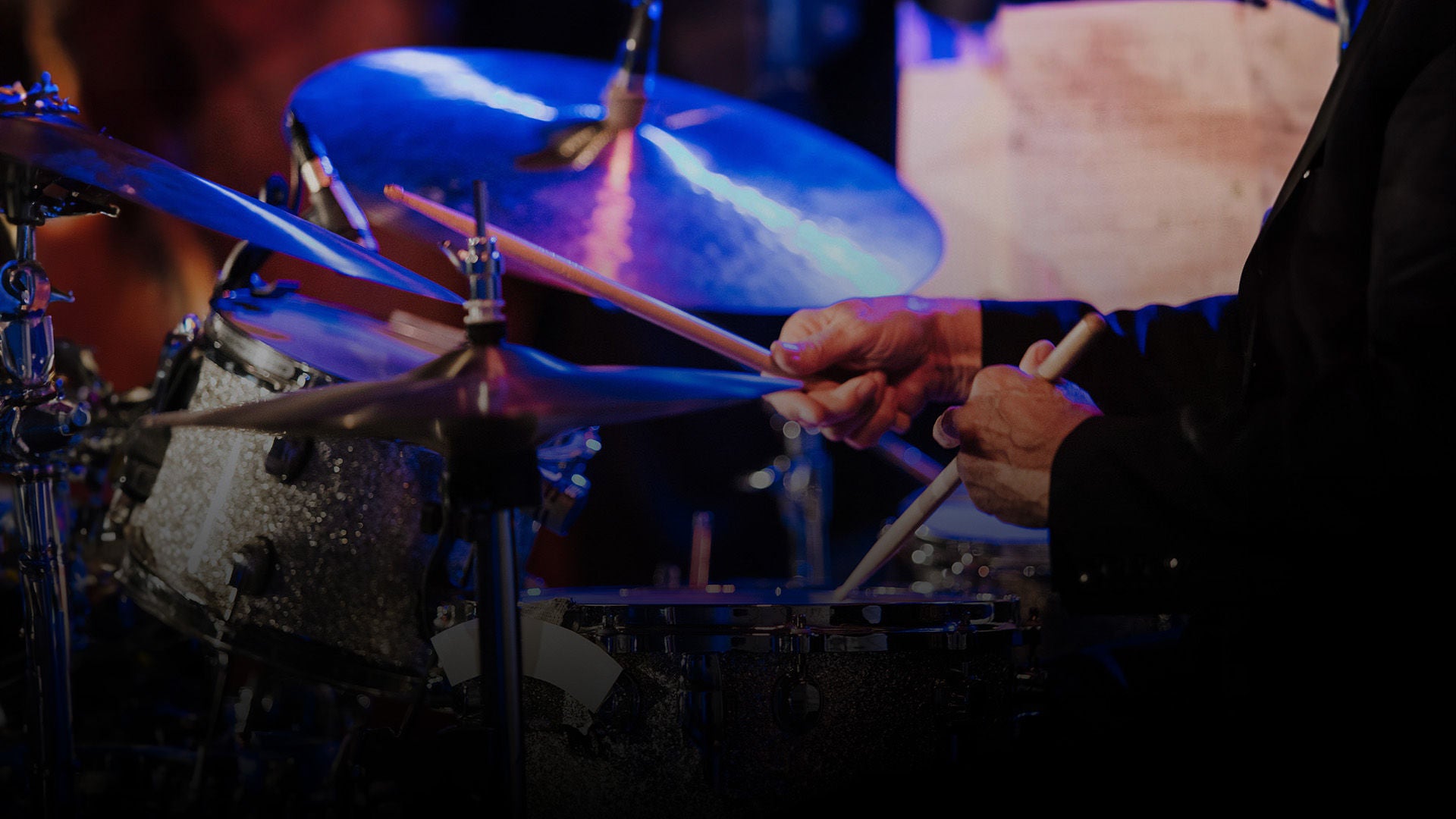 Nahaufnahme eines Schlagzeugs; Hände mit Sticks spielen auf Snare und Becken im blauen Bühnenlicht.