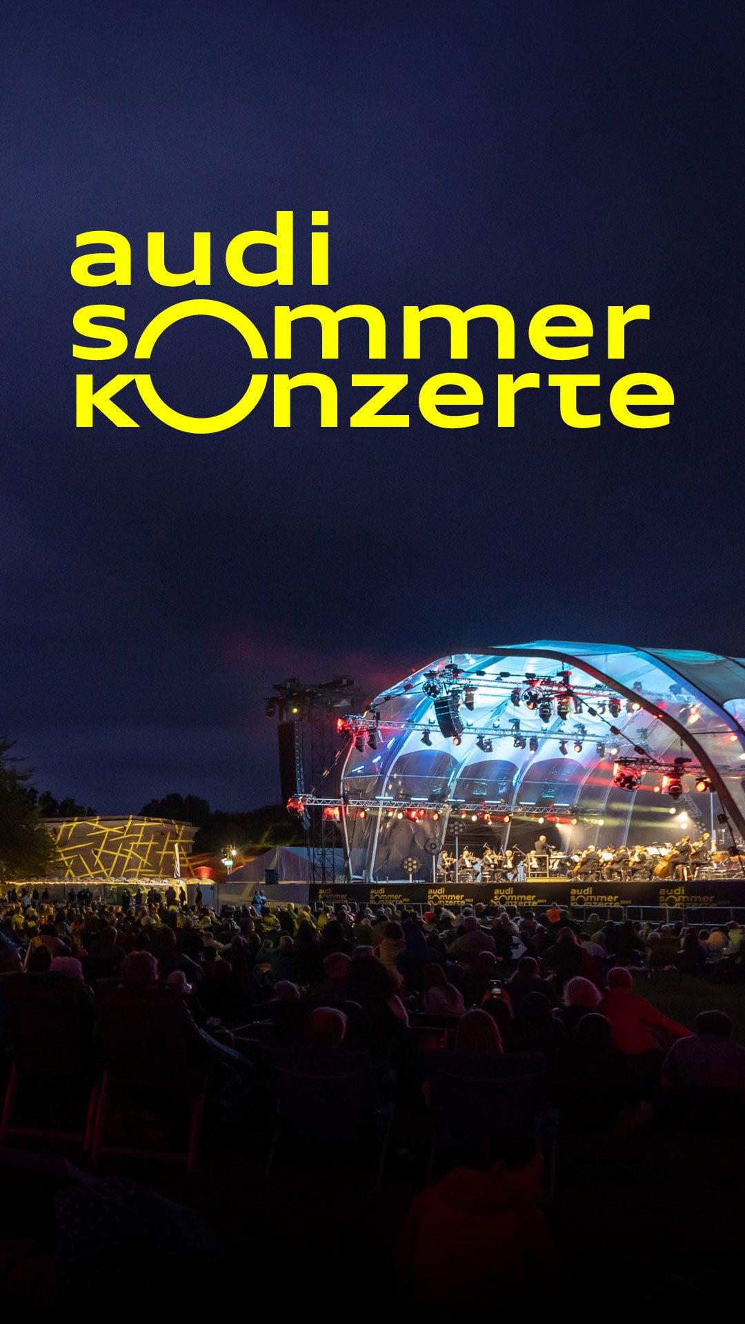 Hell beleuchtete Freiluftbühne am Abend, darüber gelber Schriftzug: Audi Sommerkonzerte.