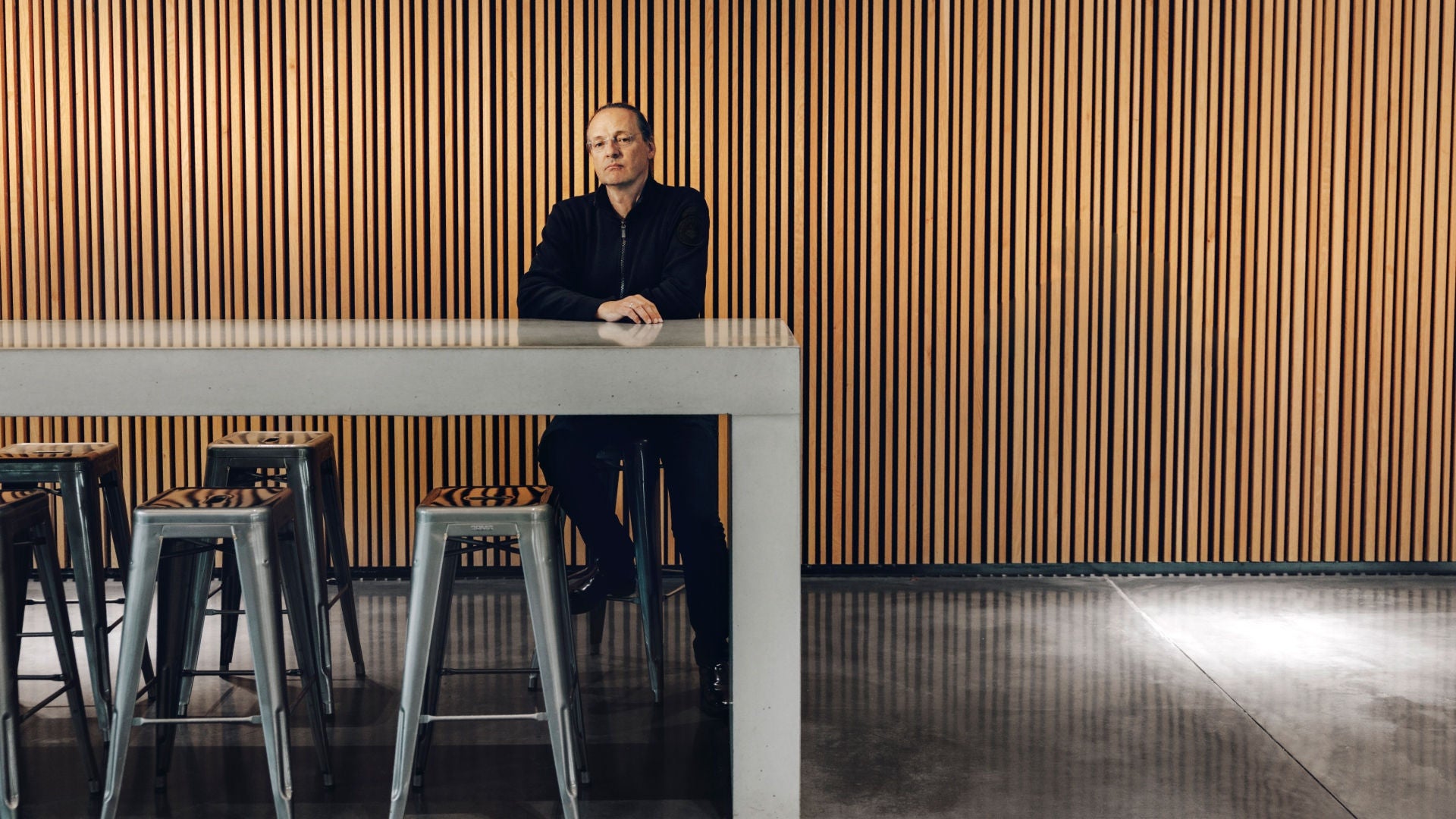 Mann sitzt allein an hohem Tisch mit Hockern vor Wand mit senkrechten Holzlamellen.