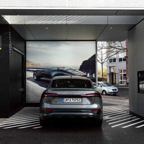 Ein grauer Audi Q8 e-tron lädt an Station 04 einer überdachten Ladesäule, im Hintergrund ein Bild eines Konzeptfahrzeugs.