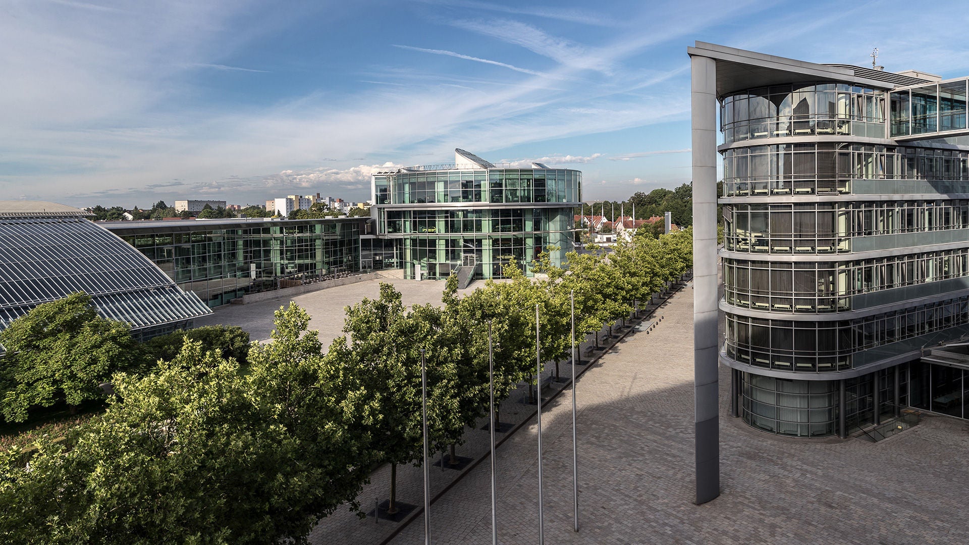 Außenansicht eines modernen Gebäudekomplexes (Audi Forum Ingolstadt) mit Glasbauten, Innenhof, Bäumen und freiem Platz.