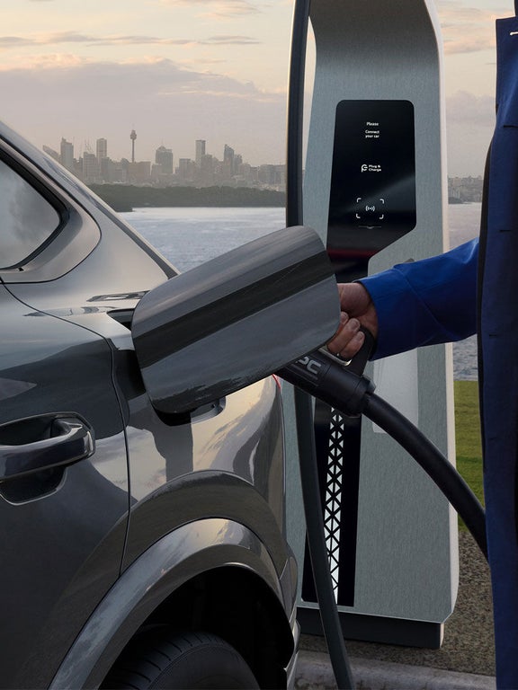 Ein Mann mit blauem Mantel hält den Ladestecker am Anschluss eines grauen Audi Q6 Sportback e-tron, im Hintergrund Wasser und Skyline.