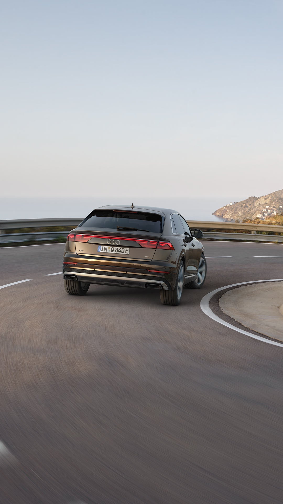 Ein brauner Audi Q8 SUV TFSI e fährt eine Küstenstraße entlang, Blick von hinten mit Meer und Bergen im Hintergrund.