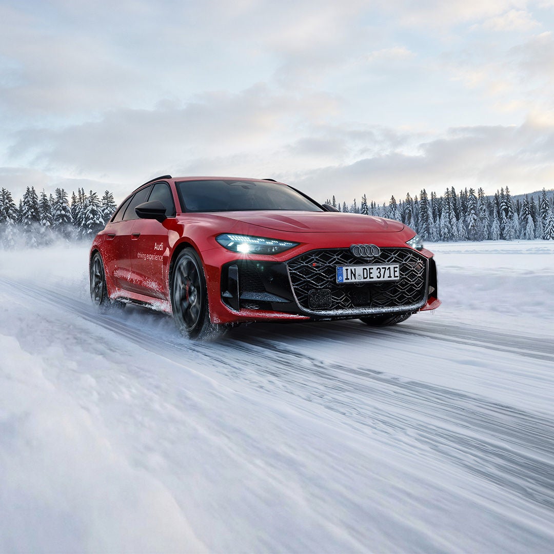 A red Audi RS 5 Avant speeds along a snow-covered road; snow flies up, with a winter forest in the background.