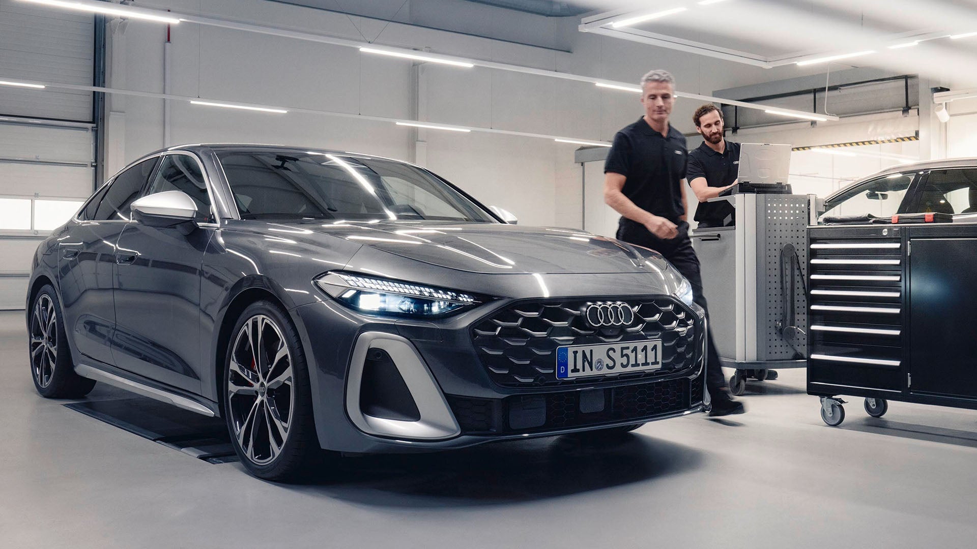 Grauer Audi steht in Werkstatt, zwei Mitarbeiter arbeiten mit Diagnosegerät daneben.