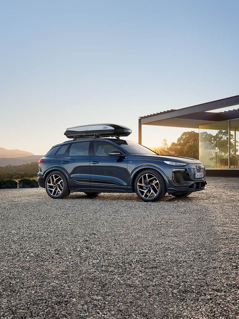 Ein blauer Audi Q6 SUV e-tron mit Dachbox vor modernem Haus, Frau auf Terrasse blickt in die Landschaft bei tief stehender Sonne.